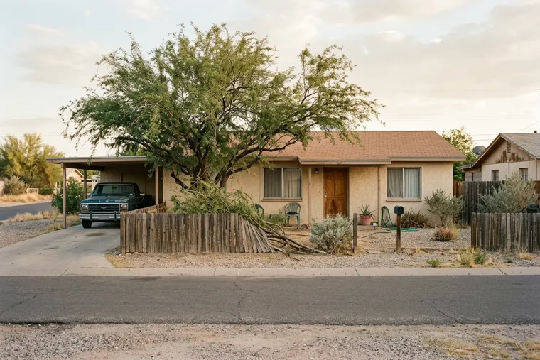 A sun-bleached single-story ranch house with a gravel yard and an overgrown mesquite tree