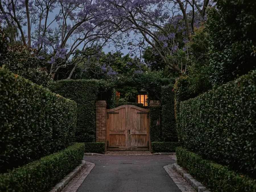 A gated driveway screened by tall hedges on a quiet residential street at dusk